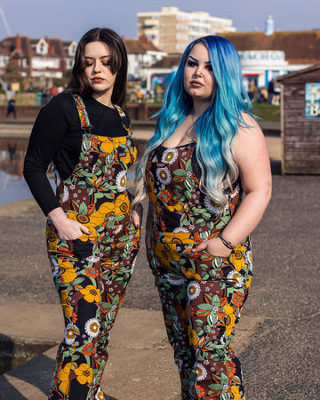 Alice and Azariah stood near a beach park area wearing the 70's black floral stretch skinny flared dungarees. They are both facing forward angled inward to highlight the flared shape. Alice has mid length brown hair and is wearing a long sleeve black tshirt underneath with black platform boots. Azariah has long blue gradient hair and is wearing a strappy black crop top underneath with black boots.