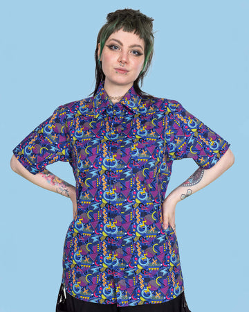 Faeryn is stood in a photography in Hove in front of a blue backdrop wearing 90's Arcade Short Sleeve Shirt with black trousers. The shirt print features classic 90's style shapes, squiggles and doodles in green, orange, pink, purple and light blue all on a dark blue background. Faeryn is posing at the camera with their hands on their hips.