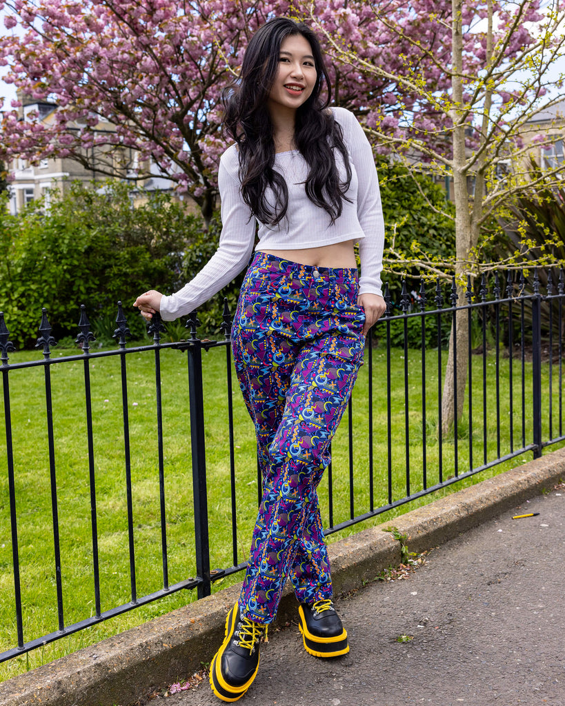 Model with long dark hair is stood outside in Hove in front of a blossom tree wearing 90's Arcade High Waist Straight Leg Jeans with a long sleeved white crop top and black and yellow boots. The jeans print features classic 90's style shapes, squiggles and doodles in green, orange, pink, purple and light blue all on a dark blue background. The model is smiling holding onto a railing with her feet crossed and one hand resting in the pocket of the jeans.