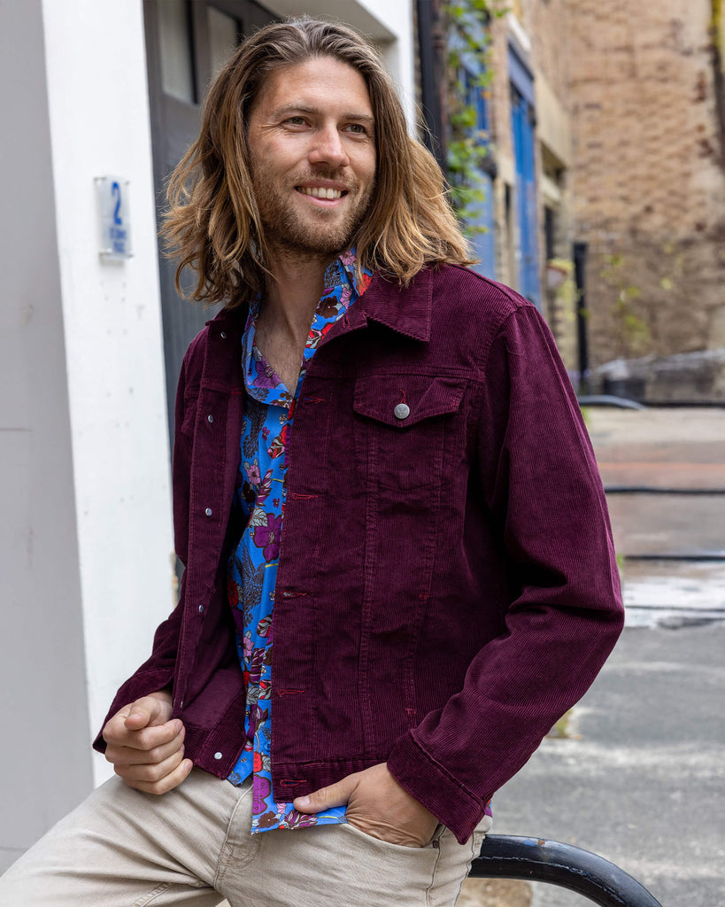 Model with shoulder length golden hair wearing Retro Vintage Burgundy Corduroy Unisex Western Jacket, paired with colourful shirt and beige trousers. The collared jacket is a burgundy colour with pockets on the front and silver buttons. The model with posing with the jacket open to reveal shirt underneath. Model is smiling with one hand in trouser pocket.