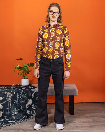 Jack is stood in a studio space wearing the Black Stone Wash Regular Rise Bell Bottom Super Flares with the longsleeve retro brown flowers shirt and white trainers. He has mid length blonde hair and round glasses, he is stood facing the camera with both arms resting by his side. The denim jeans trousers are regular rise with a stonewashed lighter black colour, fitted to thigh and flared bottoms.