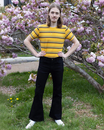 Jack is a tall blonde male with shoulder length hair looking into camera and smiling wearing a yellow retro tee and black bell bottom flares against a pink blossom tree