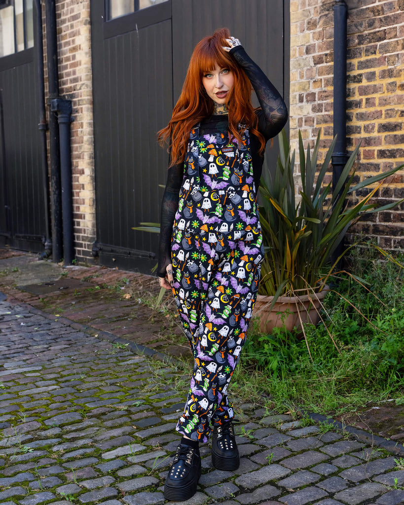 Erin Porter strikes a casual yet edgy pose in her full-length Halloween-themed dungarees by Run & Fly. She plays with her red hair, standing against a backdrop of a rustic cobbled street and tall greenery. Her outfit radiates spooky fun, featuring playful Halloween prints that make a bold fashion statement.