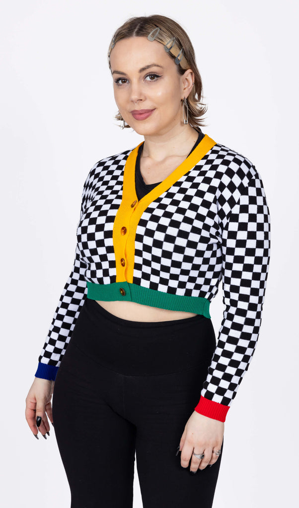 The Colour Block Checkerboard Cropped Cardigan worn by a femme model with short blonde hair with black leggings. She is facing forward smiling to camera with both hands resting by her side. The v neck crop cardigan with four brown circle buttons is a black and white checkerboard print with a yellow centre trim, a green hem trim, blue left sleeve hem and red right sleeve hem.