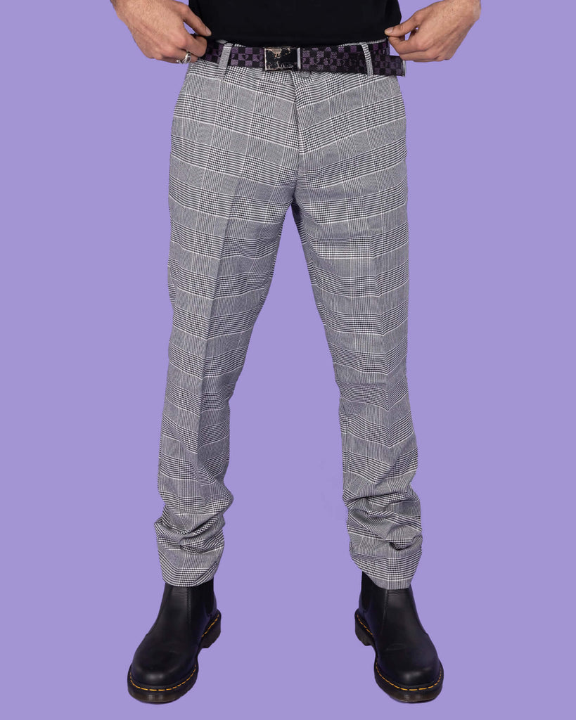 Richard, a hispanic male model with dark hair and a beard, is stood in a photography studio in front of a lilac backdrop amongst disco balls, a star shaped light and a plant wearing Retro Mod Prince Of Wales Stretch Slim Skinny Fitting Trousers with a black t shirt tucked in, a checkered belt and black boots. He is facing the camera with one leg to the side and hands behind his back.