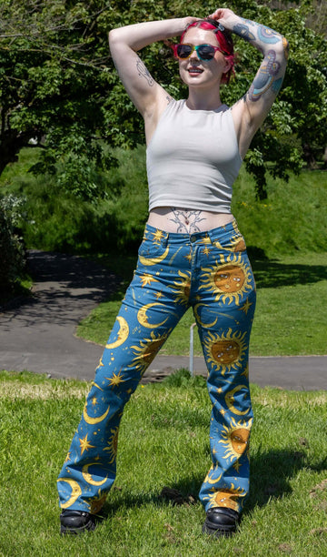 The Celestial Sun and Moon High Waisted Bell Bottom Flares shown on a femme tattooed model with red and black hair and a beige tank top, rainbow sunglasses and black trainers. She is stood outside in a grassy park area with the sun shining. She is stood facing forward with one leg bent and her arms resting above her head smiling looking off to the left. The print features gold 90s inspired moons and suns with faces, stars and sparkles all on a blue background.