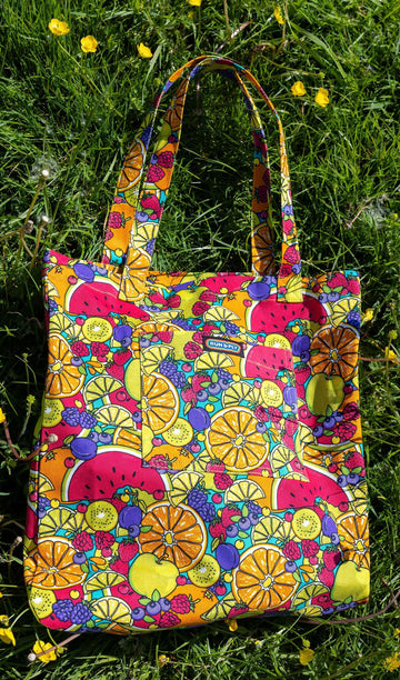 The Tutti Frutti Tote Bag laying on a grassy flowery field in the sunshine. The bright coloured tutti frutti tote bag has a front pocket with a rainbow run and fly label in the top middle. The tutti frutti print features berries, lemons, oranges, apples, watermelons, kiwis and plums all over on a blue background.