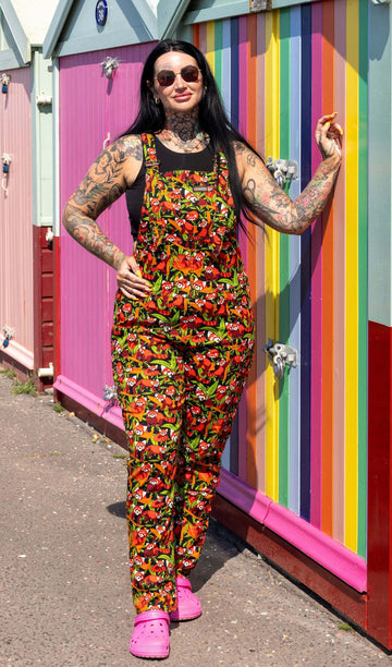 The Red Pandas Stretch Twill Dungarees worn by a femme tattooed model with long black hair, a black tank top, sunglasses and pink croc shoes. She is stood outside in the sunshine posing in front of colourful beach huts along the seafront. She is facing forward smiling with her hand on her hip and the other hand resting up on the beach hut. The black base print features various illustrated red pandas sleeping, eating and curled up on bamboo.