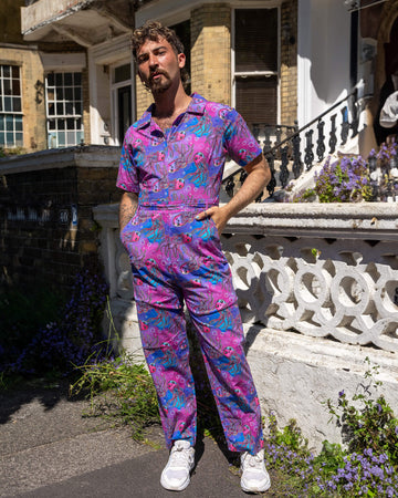 The Jellyfish Jumpsuit worn by a masc non binary person with a curly brown mullet and white trainers outside on a pavement with tall ornate houses, white concrete and brick walls and purple flowers growing along the bottom. They are facing forward smising to camera with both hands resting in the jumpsuit pockets. The blue, pink and purple base print features various smiling pink, blue and purple jellyfish and white bubble outlines.