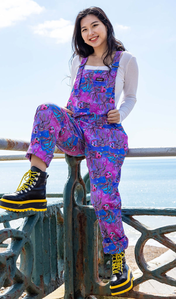 A young woman is sitting on a railing by the seaside, wearing purple jellyfish print dungarees from Run & Fly, an ethical slow fashion brand. The dungarees stand out against the backdrop of the blue sky and ocean. She pairs the outfit with black platform boots featuring yellow details, adding a bold contrast to the vibrant print.