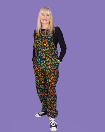 Front view of a model wearing Run & Fly bright paisley dungarees, highlighting the colourful paisley pattern. The model has long blonde hair and stands against a purple backdrop.