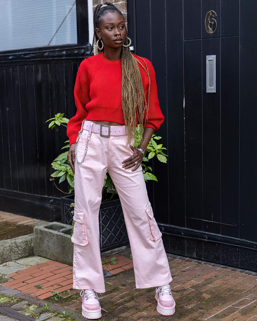 Yolande is stood outside in front of a black door wearing the 90's Baby Pink Wide Leg Oversized Stretch Cargo Trousers with a pink chain belt, red crop jumper and pink platform boots. She is facing forward leaning onto one hip with one hand in the side pocket, the other hand by her side, whilst looking off to the right. The trousers are an oversized fit, 2 flap cargo pockets on each leg as well as side pockets and back patch pockets.