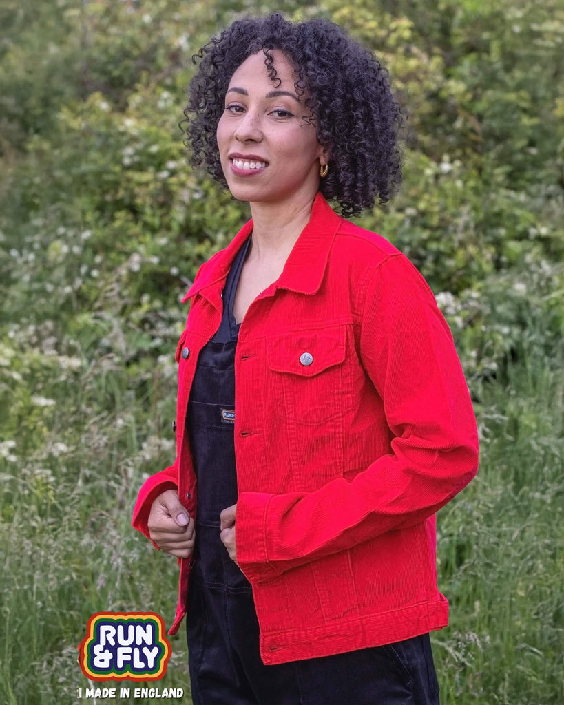 Amy is stood outside in a field area wearing the retro vintage red cord unisex western jacket with black dungarees. They are facing to the left looking over to the camera smiling whilst pulling the jacket together. Photo is cropped from the hips up.