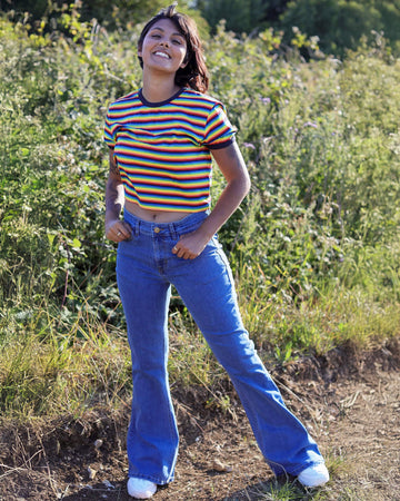 Meera is stood in a grass field wearing the stone wash blue stretch denim rock n roll bell bottom flares with a retro rainbow stripe short sleeved tshirt and white trainers. She is facing forward posing with one leg kicking forward and both hands resting on the front jean pockets whilst smising to camera.