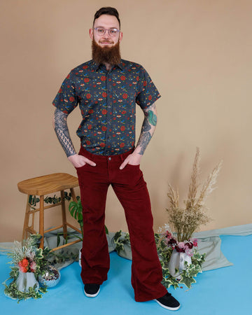 Dec, a white male with a beard and glasses, is stood in a photography studio in Hove in front of a beige backdrop wearing Tawney Port Corduroy Bell Bottom Flares with Retro Old School Tattoo Print Short Sleeve Shirt. Dec is smiling at the camera with his hands in the trouser pockets. 