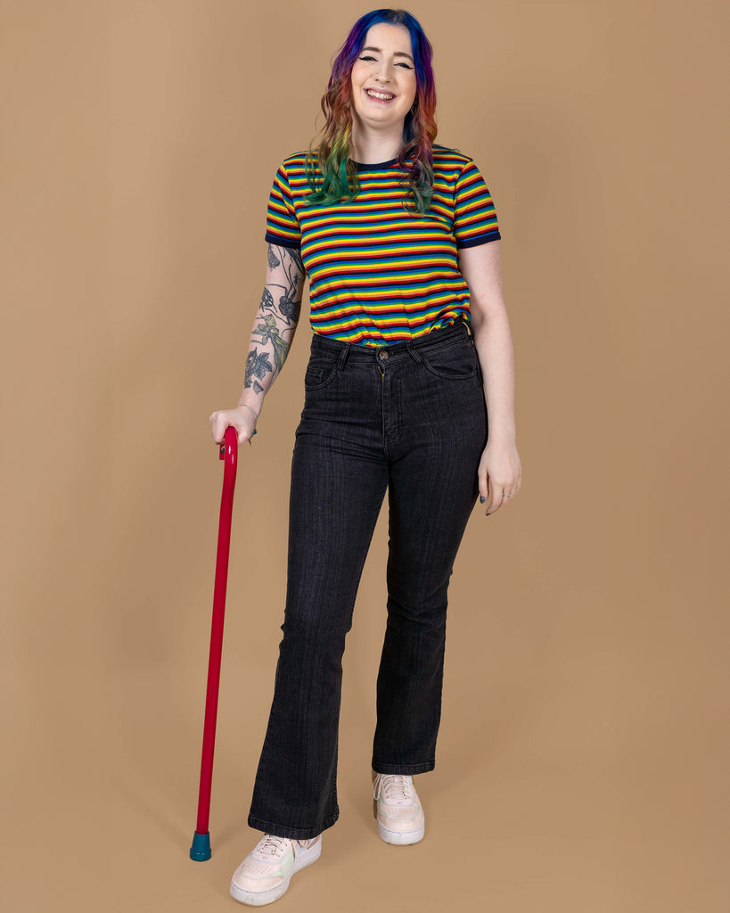 Eliza is stood wearing the Unisex Black Stonewashed Bellbottom Flares Leg High Waisted Jeans with the rainbow stripe short sleeve tshirt and pastel pink trainers. They have rainbow hair, nature themed tattoos on one arm and a red walking aid whilst smiling to camera. The jeans are high waisted with a faded stonewash black colour, fitted to thigh and flared bottoms.