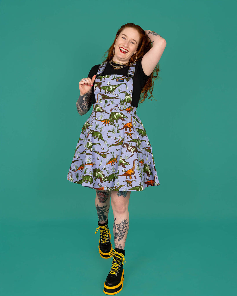 Izzy a red haired white model with tattoos is smiling wearing a blue dinosaur pinafore, black tee and black and yellow boots shot in studio against a green background. The print is a soft blue base with various dinosaurs and prehistoric plants repeating all over.