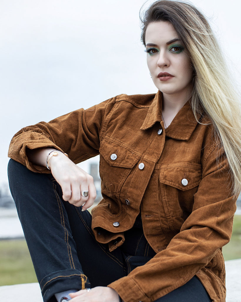 Alice with long blonde dyed hair is posing outside sat on a brick wall wearing the Retro Vintage Washed Tan Corduroy Unisex Western Jacket with blue denim jeans. She has green eye makeup and brown lipstick. She is sat leaning back with one leg up on the wall whilst smising looking down to camera.