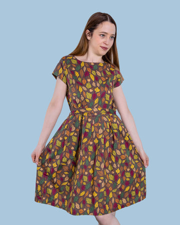 The Autumn Leaves Stretch Belted Tea Dress with Pockets on a femme model with long brown hair and black heeled boots. She is posing with one leg crossed over holding out the skirt smiling looking right at a picnic setup with a picnic basket, fruit, flowers and katie abey mugs. The dress print is a brown base with green, red, orange and yellow autumnal leaves and acorns.