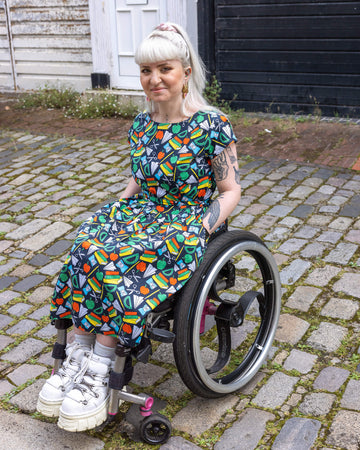 Female model seated in a wheelchair wearing Back to School Stretch Belted Tea Dress with Pockets. The print has an array of educational motifs, including books, apples, rulers, and scissors, all in bright, eye-catching colours on a navy background. 