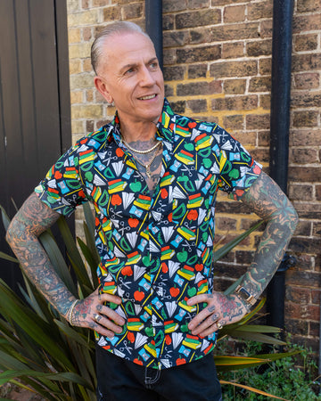 An older man with tattoos models a vibrant short-sleeve shirt featuring a pattern of school supplies on a dark base colour. He stands against a brick wall, smiling and looking to the side with his hands on his hips.