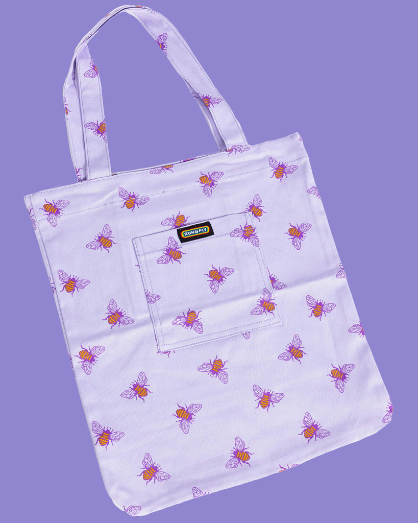 flat lay of a lilac tote bag with an all over print of yellow bumble bees with purple wings on a purple backdrop. There is a front pocket on the tote bag with Run&Fly logo on the front.