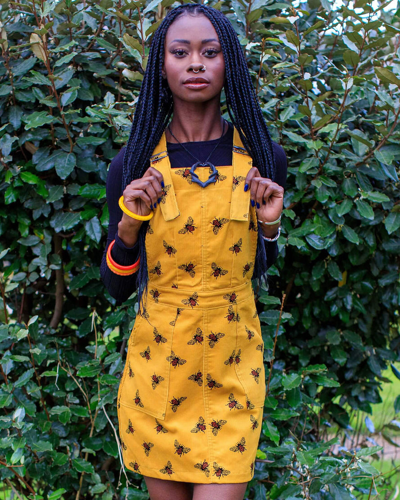 Carella is stood in front of a bush wearing the mustard corduroy the bees kneez bee print pinafore dress with a long sleeve black tshirt underneath and black boots. They are facing the camera holding onto the top of the pinafore bib whilst smising.