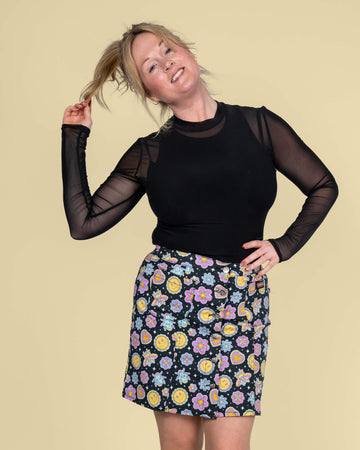 Karina playfully poses in the Run & Fly mini skirt designed by Waffle Mama Design. The black skirt is decorated with whimsical smiley faces, hearts, and flowers, exuding retro fun against the soft beige background.