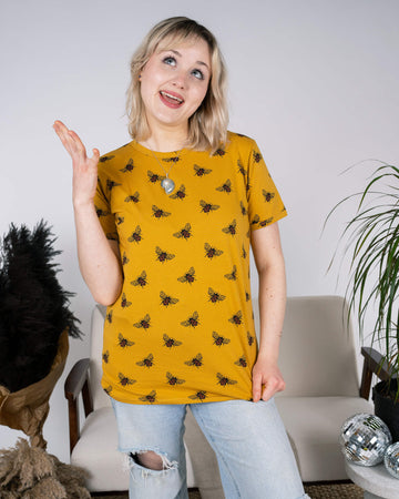 A playful shot of a person in the Run & Fly Buzzy Bee Mustard Gold Tee, smiling and gesturing with one hand. The mustard yellow t-shirt features a charming bee print, styled with light-wash ripped jeans. The setup includes a neutral interior with plants and disco balls, reflecting the cheerful vibe of this ethical, small-batch slow fashion piece.