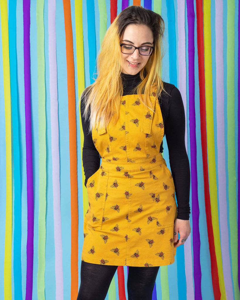 Jojo is stood in front of a blue rainbow studio background wearing the bees knees bee print gold stretch twill pinafore dress with a long sleeve black tshirt and grey boots. She is facing forward leaning back on one leg with one hand in the pinafore pocket whilst smiling and looking down.