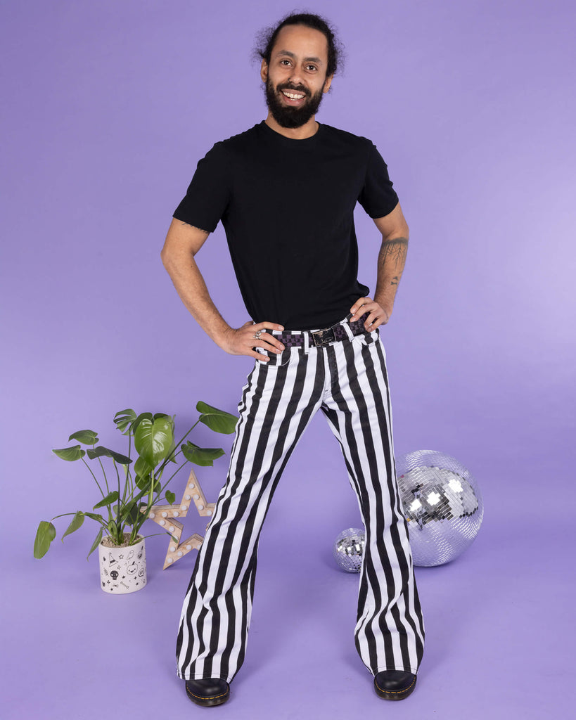 Richard, a hispanic male model with dark hair in a bun and a beard, is stood in a photography studio in front of a lilac backdrop wearing Black & White Striped Bell Bottom Stretch Super Flares with a black t shirt, black and purple checkered belt and black shoes. Richard is smiling and facing the camera with his hands on his hips. Run & Fly logo is at the corner or the photo with 'Made in England' written underneath.