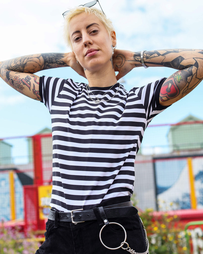  YO poses confidently in a black-and-white striped T-shirt, accessorised with black trousers featuring a statement O-ring and chain detail. The outdoor backdrop includes vibrant colours and street art.