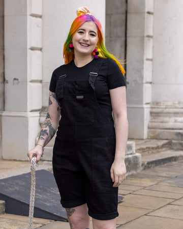 Eliza, a non binary femme person with rainbow hair using a clear sparkly walking stick, smiling and wearing the Black Denim Stretch Dungaree Shorts with a black tee underneath and multicolour pompom earrings. They are stood outside in a cobble street in london posing facing forward.