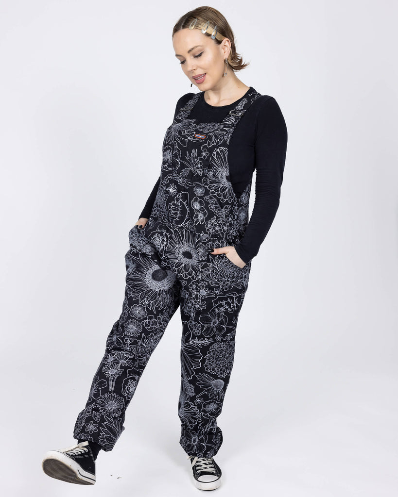 The Black and White Floral Stretch Twill Dungarees worn by a femme model with short blonde hair with a black long sleeve top and black trainers on a white background. She is facing forward with both hands in the dungaree pockets looking down kicking one leg forward. The dungaree print is a black base with various floral and botanical plants in a white outline.