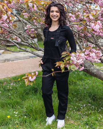 Sophie with mid length brown hair smiling by a pink blossom tree wearing the Black Stretch Denim Dungarees with a long sleeve black t-shirt and white trainers. 