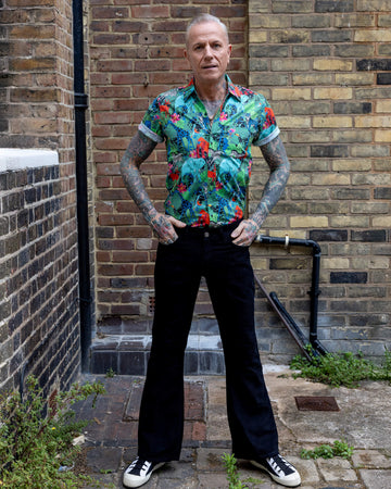 Jim is standing in a mews in Hove he is a tattooed grey haired man he is wearing a jungle print shirt and black flares by Run & Fly