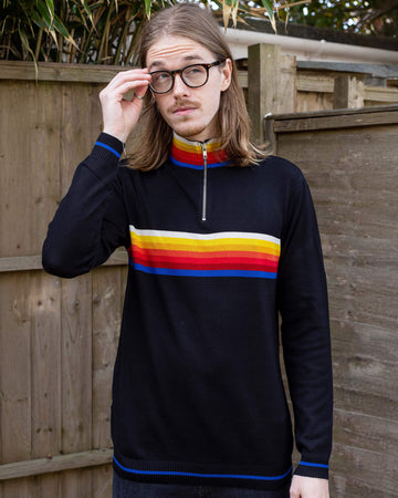 Jack is a man in his 20's with shoulder length hair and a little beard wearing glasses and a retro knit high neck Run & Fly jumper with striped detail on the front. He is stood outside against a fence adjusting his glasses.