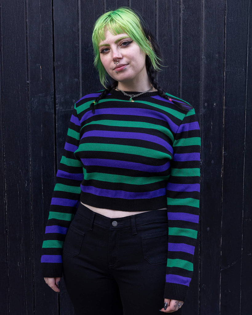 Model with green and black hair wearing Black Striped Purple and Green Cropped Jumper paired with black jeans. The jumper is a cropped style and has a black background colour with chunky muted green and purple horizontal stripes across it. Model is smiling at camera.