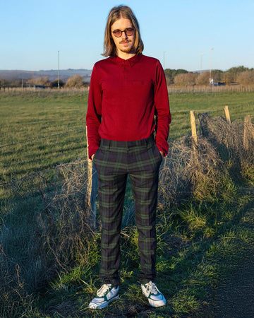 Jack is stood in front of a grass field wearing the retro mod stretch black watch tartan trousers with a long sleeve red shirt and white trainers. They are facing the camera posing with both hands in their trouser pockets and smiling to camera.