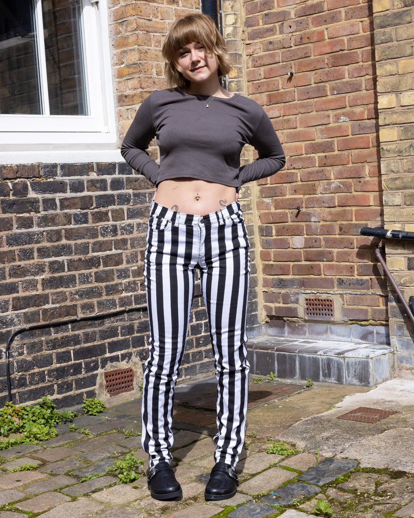 Julia is a white woman in her early 20's wearing black and white striped mod jeans with a charcoal top and black slide on shoes stood by a brick wall