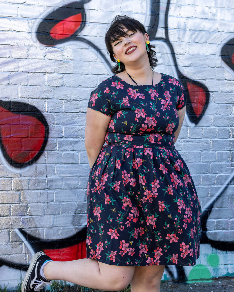 Rebecca is stood in front of a graffiti wall wearing the cherry blossom black belted stretch tea dress with pockets with black trainers. She is facing forward posing standing on one leg with her arms crossed behind her and smiling to camera.