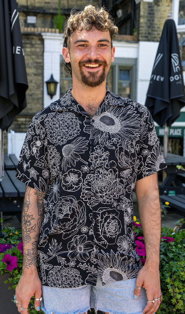 The Black and White Floral Short Sleeve Rayon Shirt worn by a masc non binary person with a brown curly mullet and denim shorts. They are stood outside in front of a pub garden facing forward laughing with both hands resting by their side. The black base shirt features various floral outlines in white with a button up front and slight v neck collar.