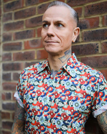 Jim is stood in front of a brick wall wearing the red and blue record short sleeve shirt with blue denim jeans. They are heavily tattooed with swept back silver hair. They are facing the camera posing with one hand on their hip whilst the other rests by their side. Photo is cropped from the hips up.