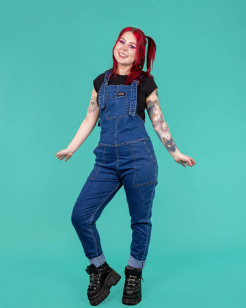 Flo a red haired femme white model with tattoos is wearing blue denim dungarees with a black tee and black platform trainers shot in a studio by Amy Davies Photography