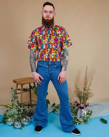 Declan a man in his 20's with a beard, tattoos and glasses is wearing a pair of blue denim flares with a shirt with all over dog print on and is standing against a beige background with a stool, florals and mirrorball