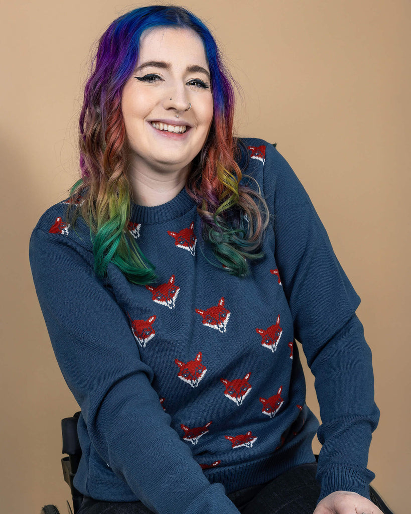 Eliza with rainbow hair is sat in their wheelchair wearing the blue fox head jumper with black stone wash flares. They are leaning forward with both hands resting on their lap and smiling to camera. The blue fox head jumper is a petrol blue base colour with red and white fox heads repeating over the middle of the jumper segment, the arms and back are plain petrol blue.