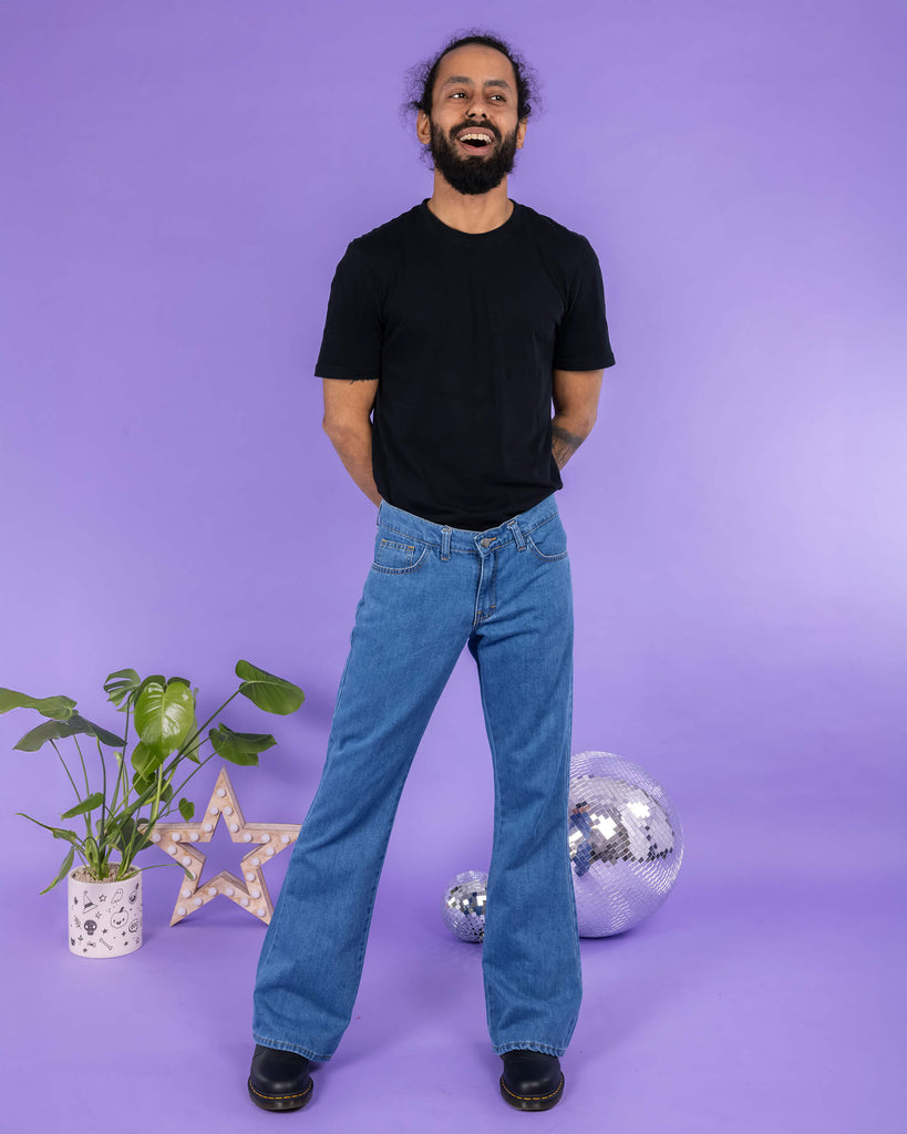 Richard, a hispanic male model with dark hair in a bun and a beard, is stood in a photography studio in Hove in front of a lilac backdrop wearing Stonewash Retro Boot Cut Flare Jeans with a black t shirt and black shoes. Richard is smiling and facing the camera with his arms behind his back and legs slightly apart. Run & Fly logo is at the corner or the photo with 'Made in England' written underneath.