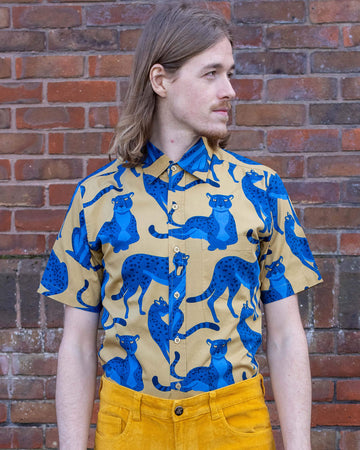 Jack is stood in front of a brick wall wearing the blue leopard short sleeve shirt with gold corduroy flares and tan boots. They are facing forward posing with both arms resting by their side whilst looking off to the right.