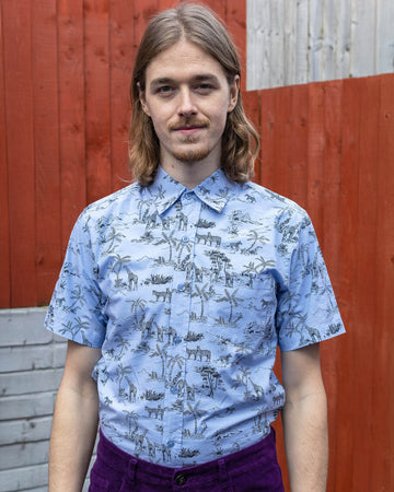 Jack is stood in front of a brick and wood panelled wall wearing the blue safari short sleeve shirt with purple corduroy flares. They are facing forward posing with both arms by their sides whilst smiling to camera. Photo is cropped from the hips up.