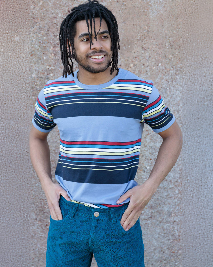 Richard, a hispanic male model with dark hair in a bun and a beard, is stood in a photography studio in Hove in front of a lilac backdrop wearing Vintage Blue Stripe Tee with blue jeans. The blue t shirt has horizontal lines in red, blue, white, yellow and navy. Richard is facing the camera smiling and is holding the bottom of the t shirt with one hand and other resting down by his side. The photo is cropped at the thighs.